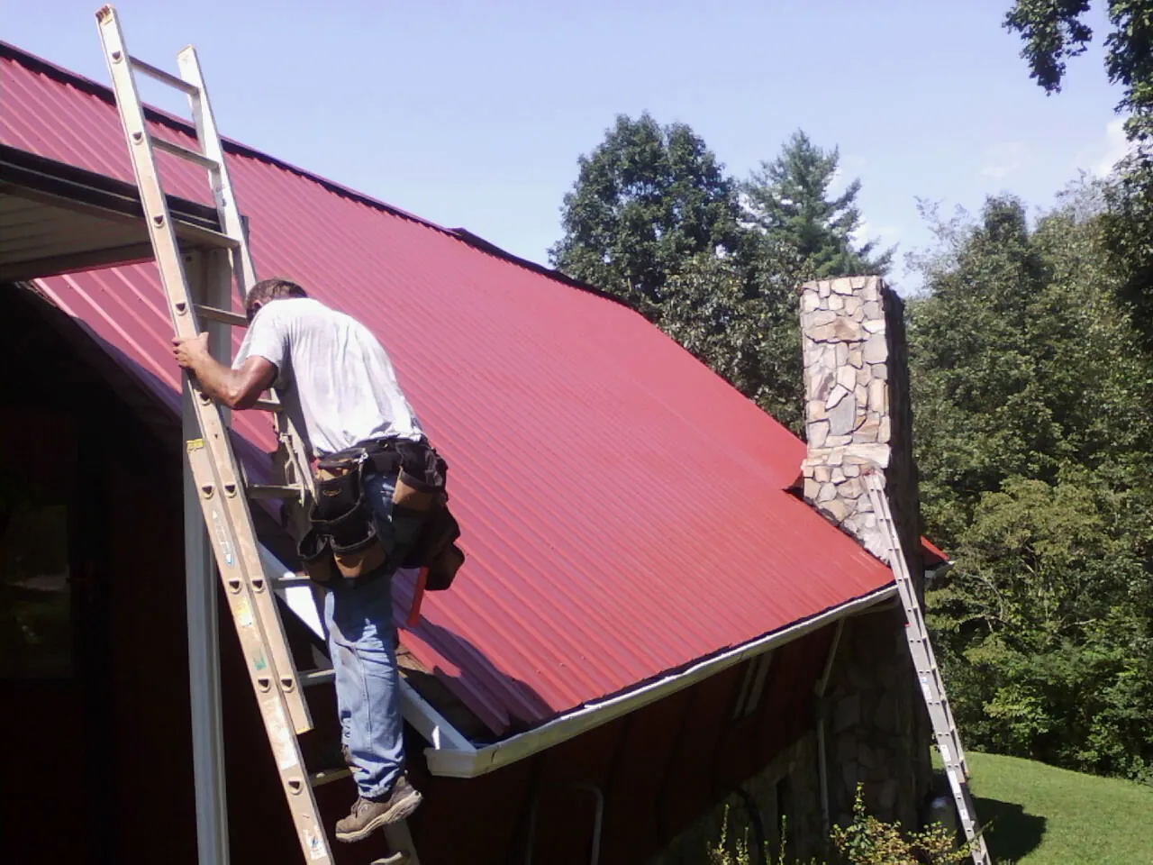 Roofer on a ladder beside a red metal roof and stone chimney — Soffit & Fascia Repair by Roofing Brothers in Greenfield Park
